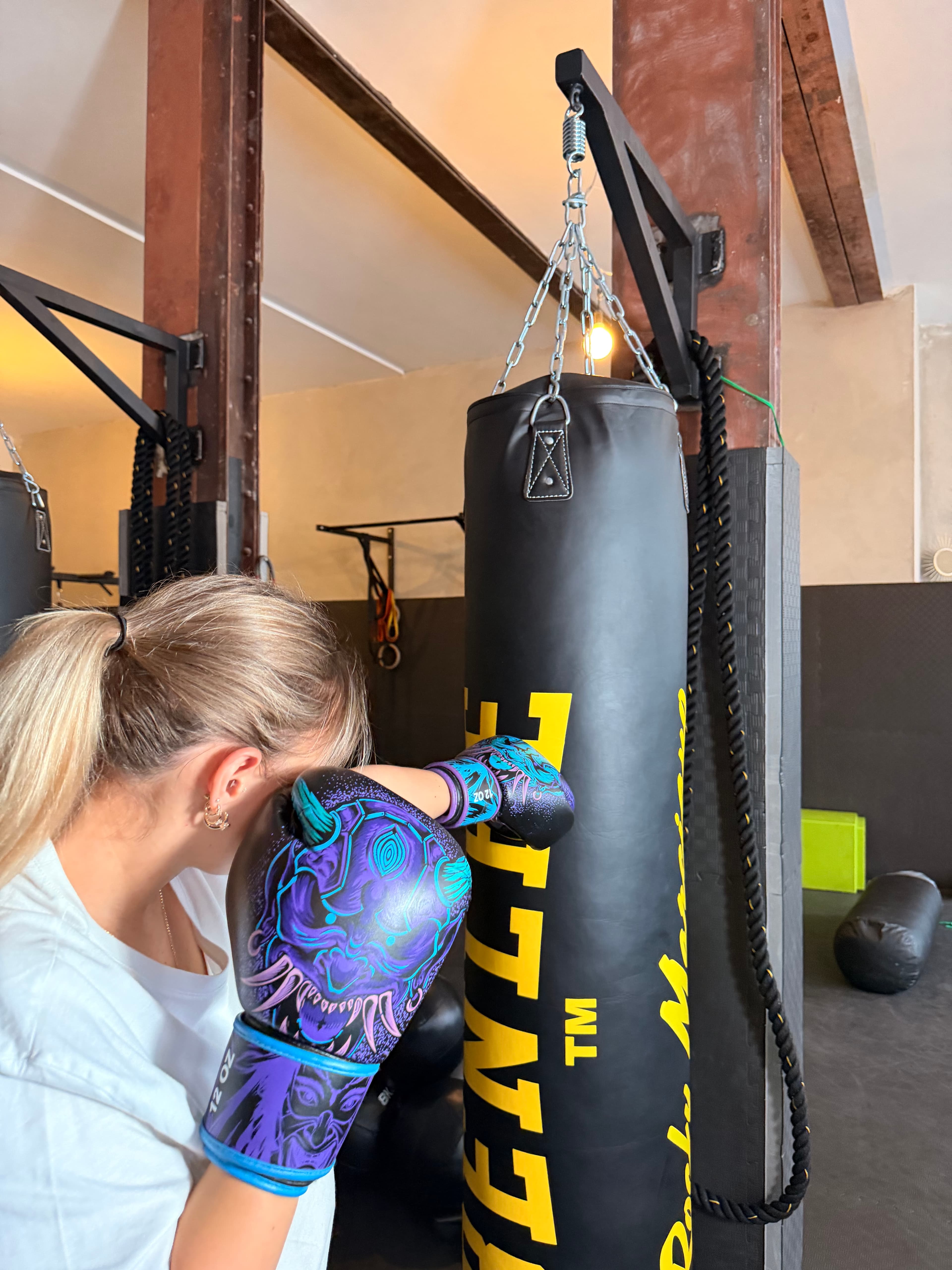 Blonde Frau mit lila-blauen Boxhandschuhen schlägt gegen einen schwarzen Boxsack in einem industriellen Boxstudio.